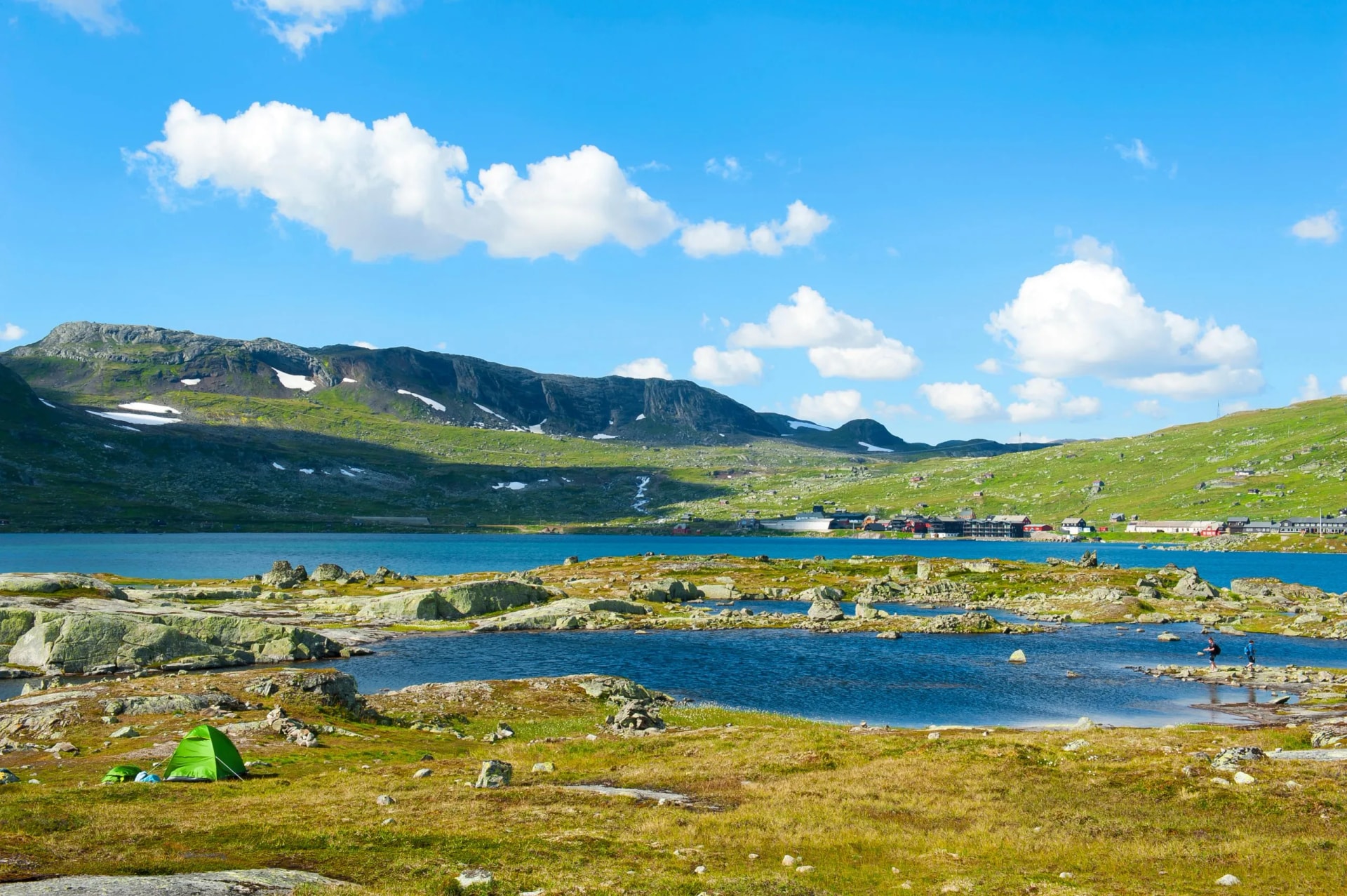 Green tent camping near rocky shore of lake with mountains and Finse village in background.