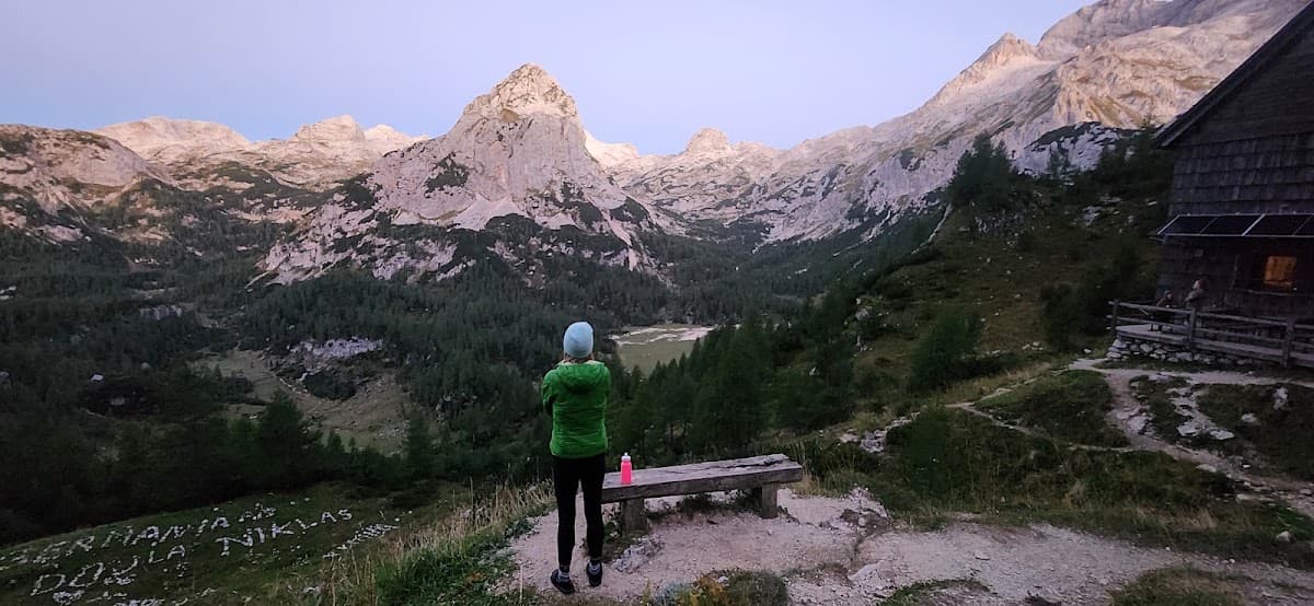 Hiker standing by wooden bench overlooking alpine mountain range and rustic cabin