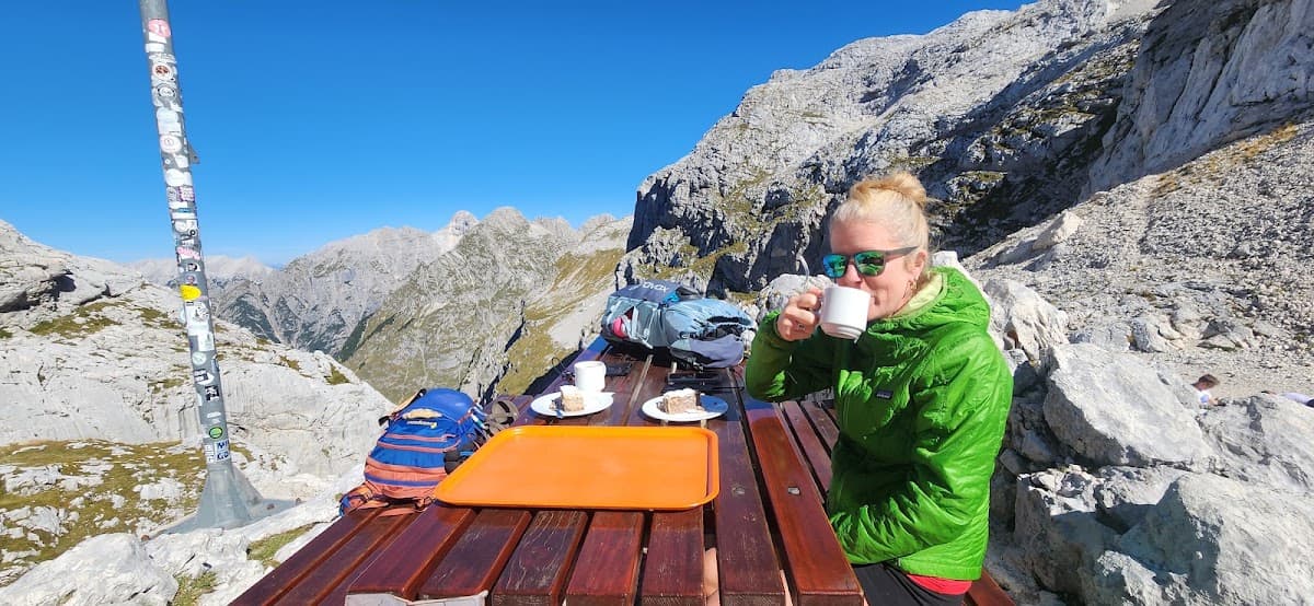 Hiker drinking from a cup at a wooden picnic table with mountain peaks in the background