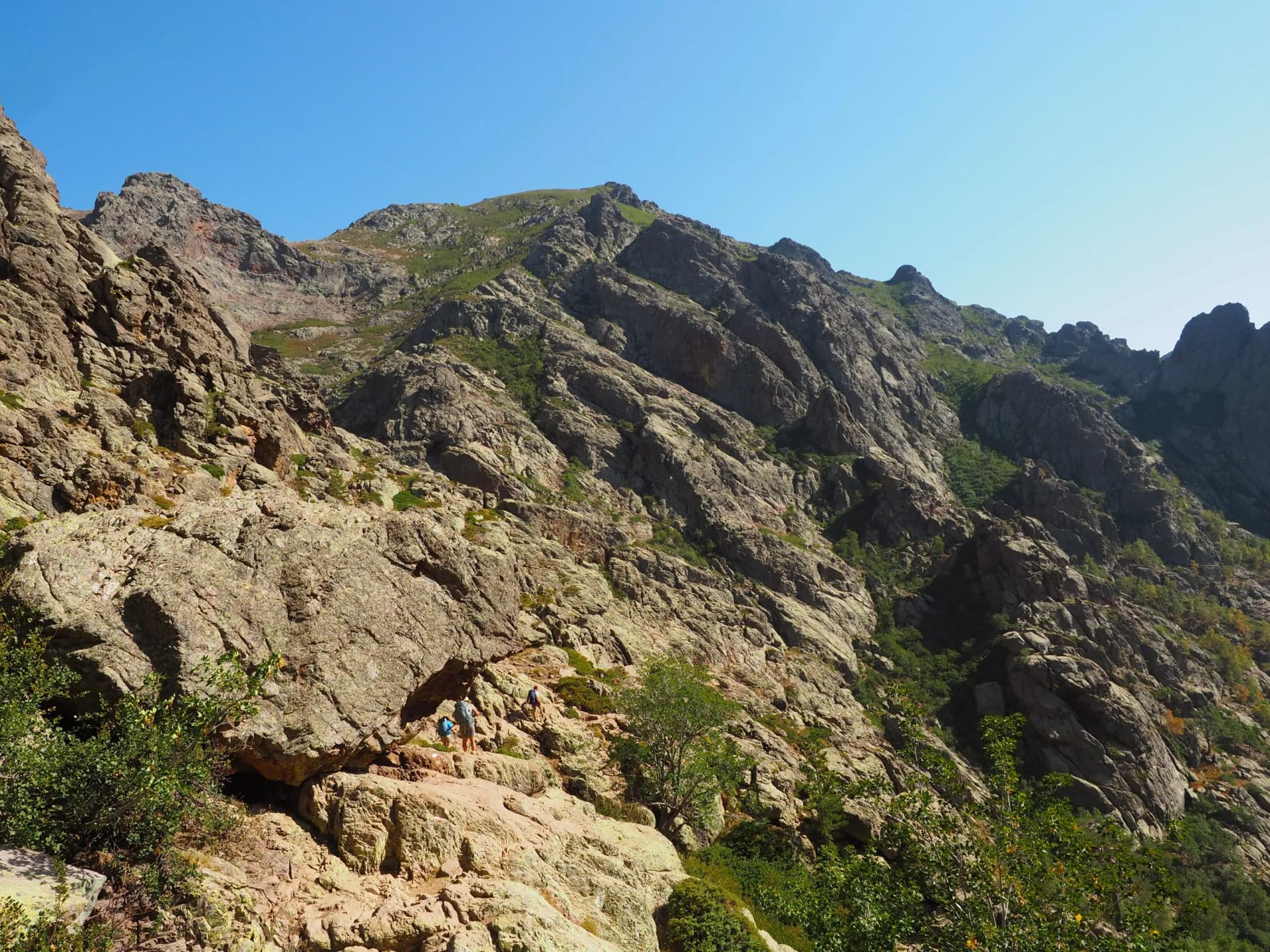 Hikers ascending rocky mountain terrain under a clear blue sky on the GR20 trail in Corsica.