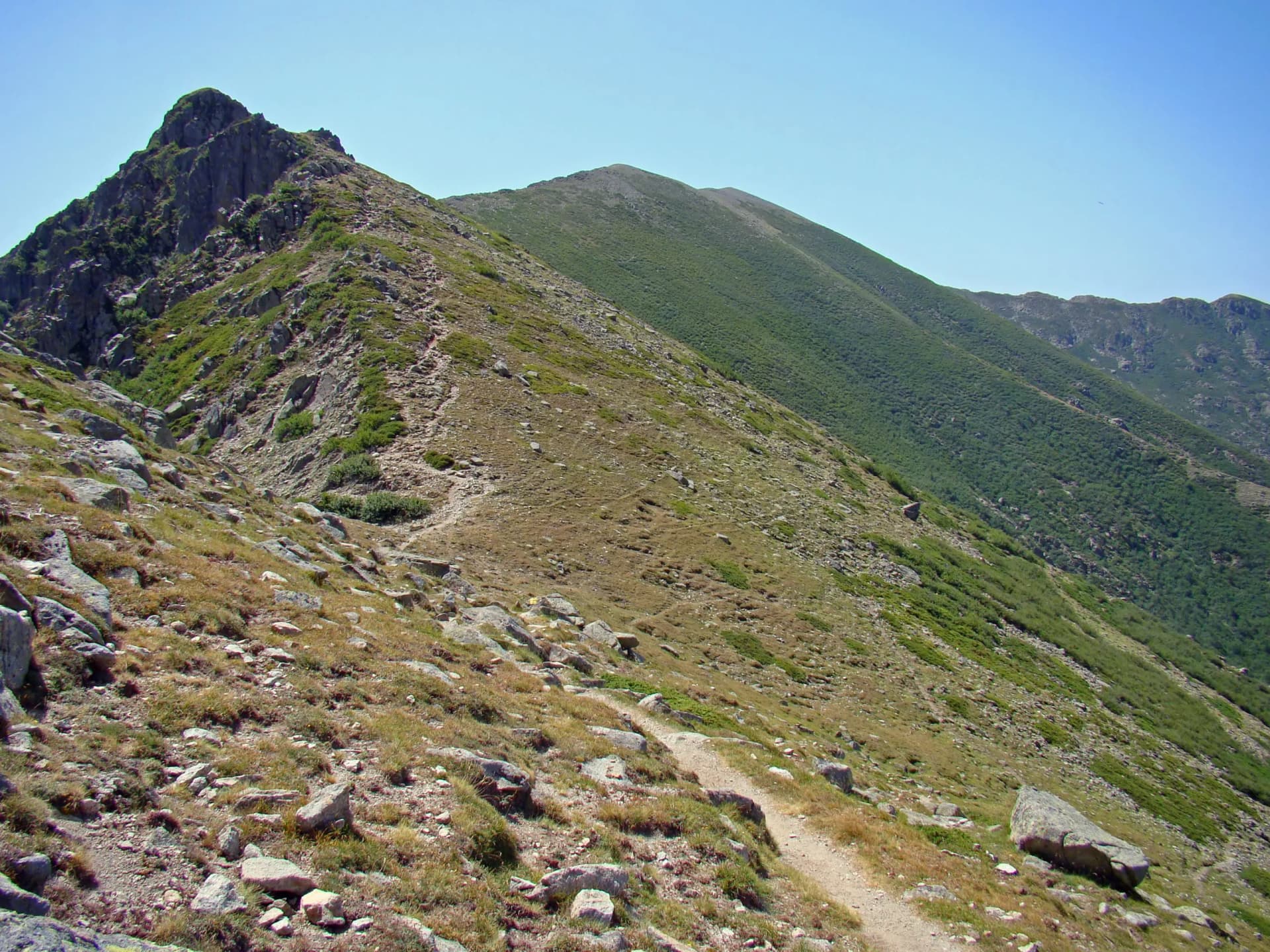 Hiking trail ascending rocky, grassy mountain slope under clear blue sky, Corsica.