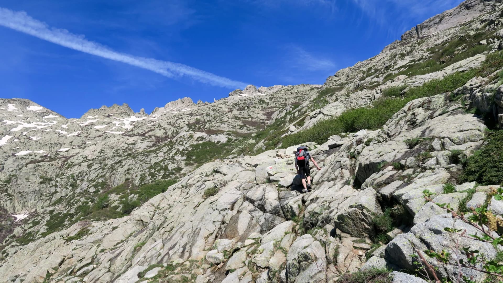 Hiker with backpack scrambling up steep, rocky terrain under a bright blue sky, GR20.