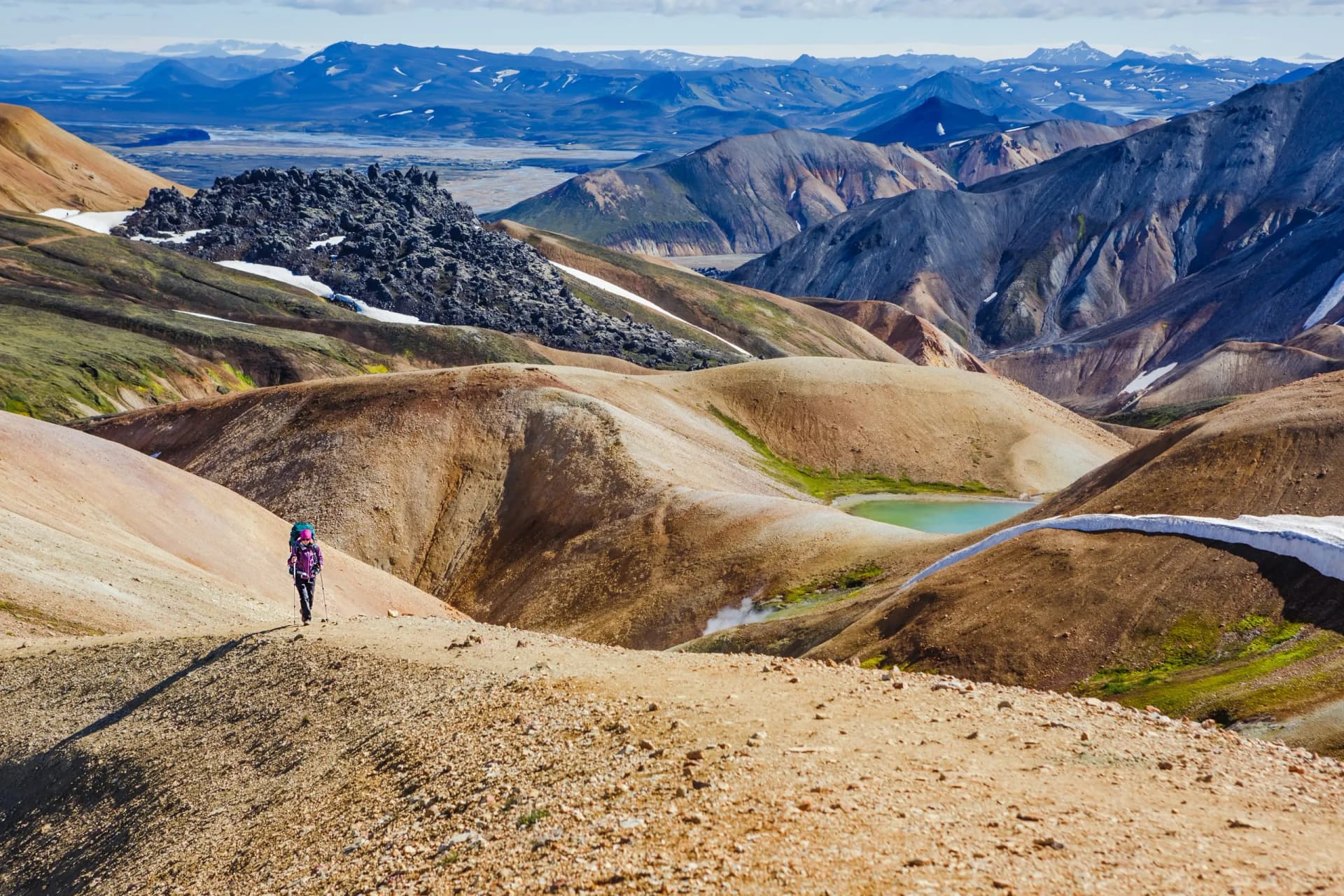 Hiker trekking through colorful, rolling hills with snow patches in Landmannalaugar, Iceland.