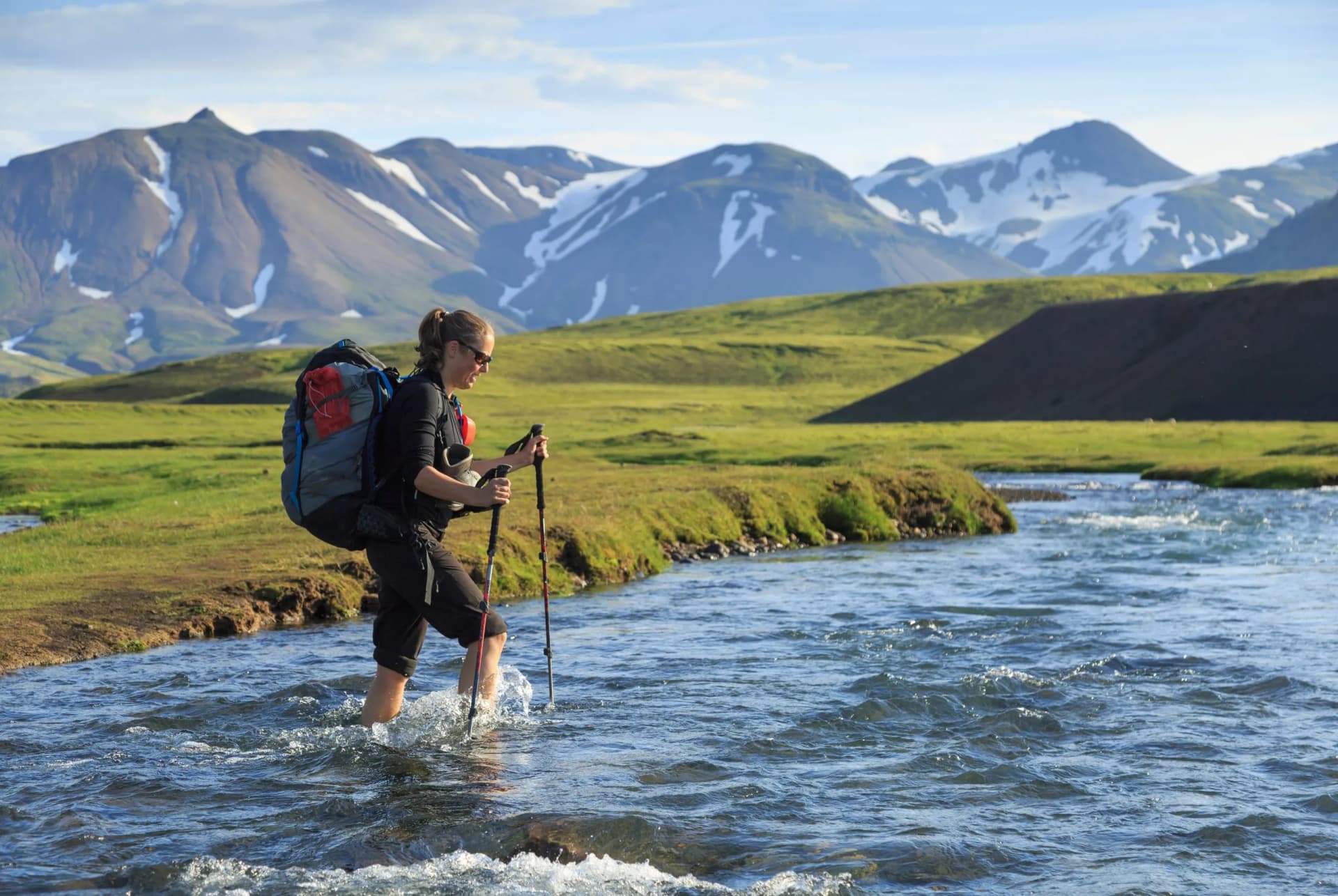 Female hiker crosses a river with trekking poles on the Laugavegur Trail, Iceland, with snowy mountains.