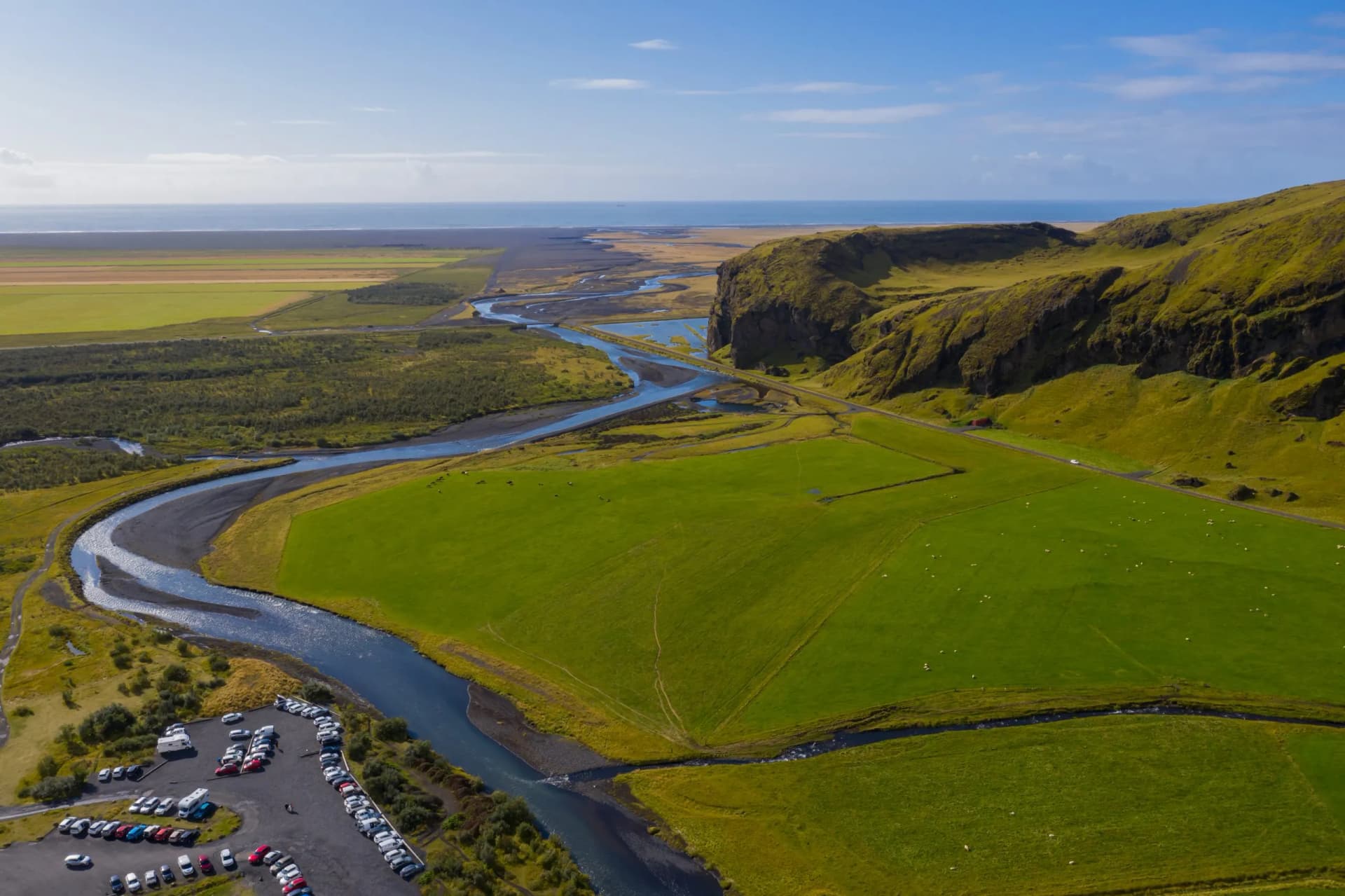 Lush green Skoga River valley near Skogafoss waterfall with parking lot and ocean view.