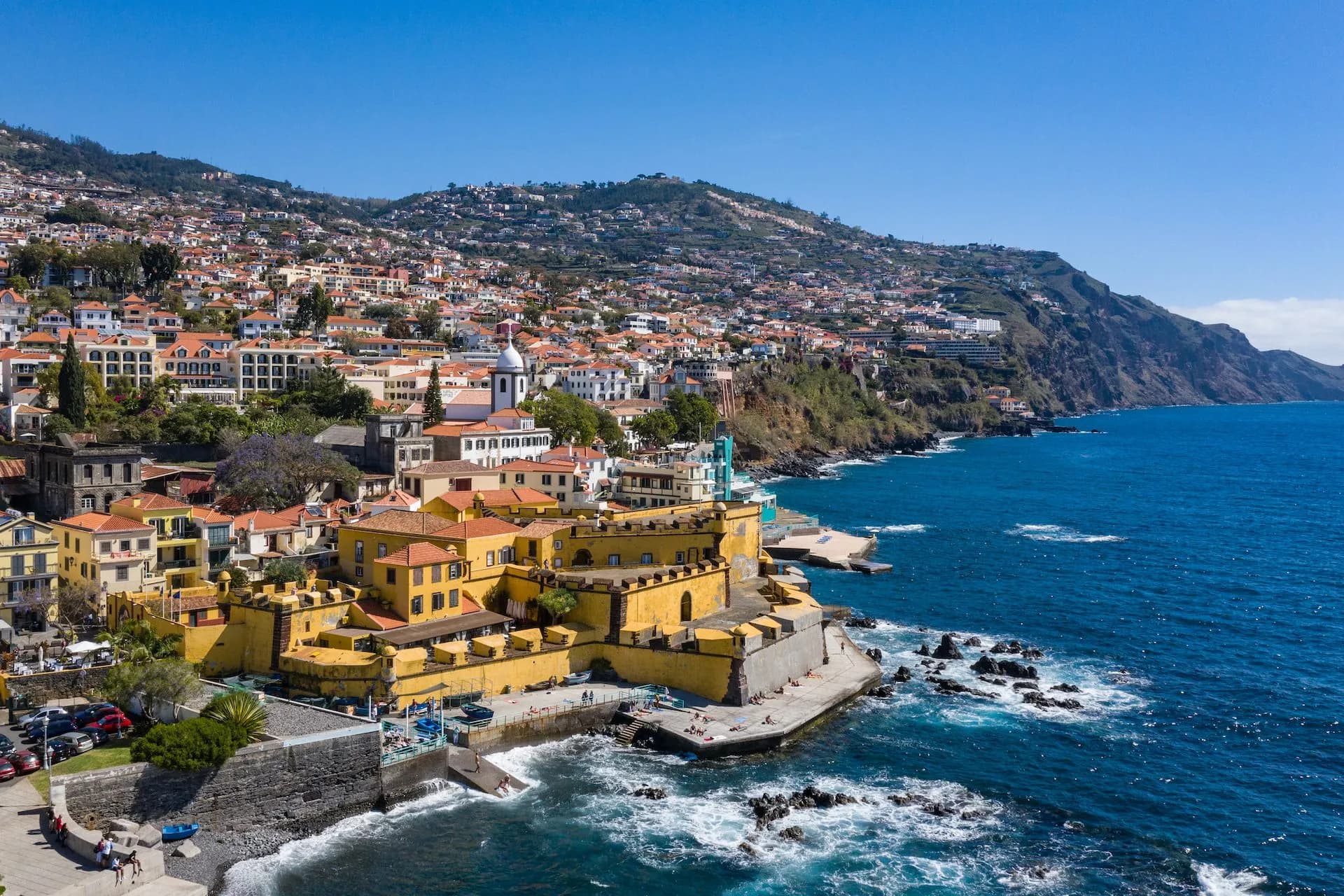 Coastal city of Funchal with yellow fortress on the water and steep hillsides under blue sky.