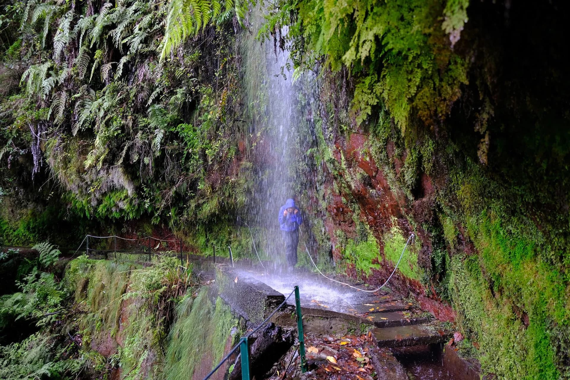 Hiker in blue jacket standing under waterfall on mossy levada path in Levada do Rei.