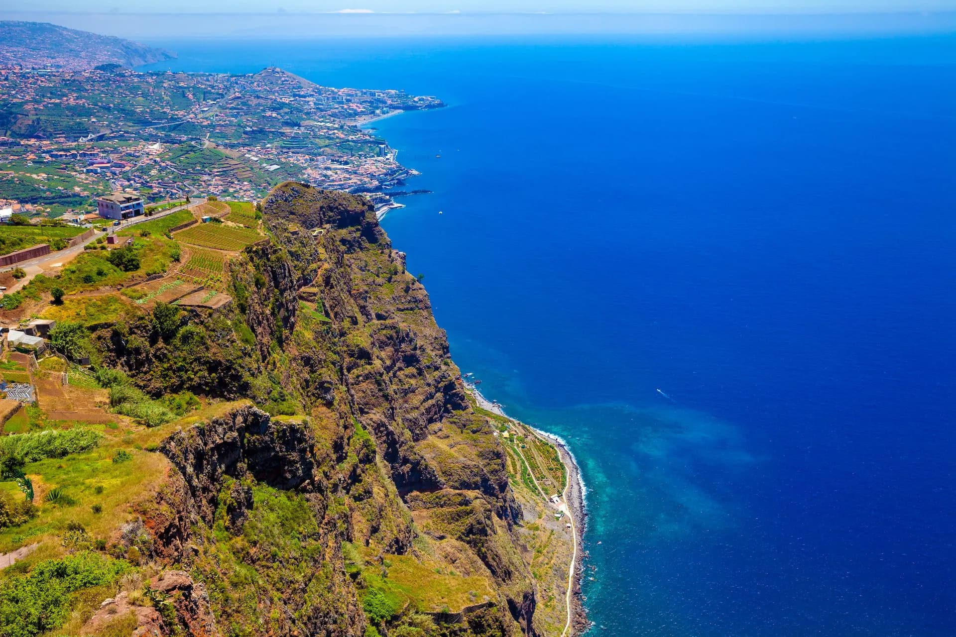 View from Cabo Girao cliff overlooking steep green terraces, coastal road, and deep blue Atlantic Ocean.