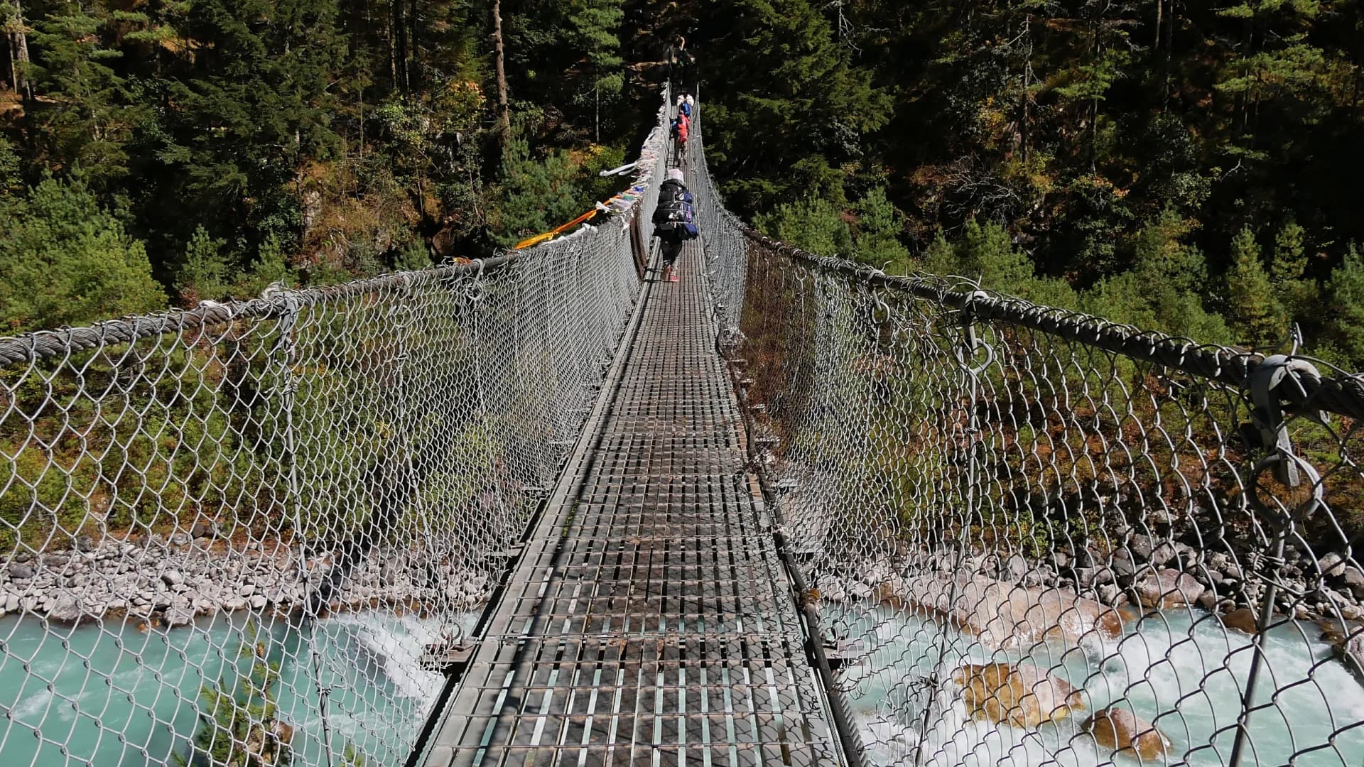 Porters crossing a suspension bridge with attached Buddhist prayer flags above Dudhkoshi River in a valley near Manjo on Mount Everest Base Camp Trek, Nepal.