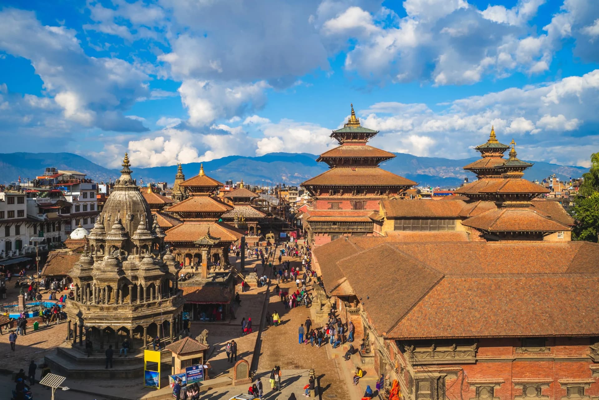 Patan Durbar Square in Kathmandu, Nepal, with tiered temples and crowds under a blue, cloudy sky.