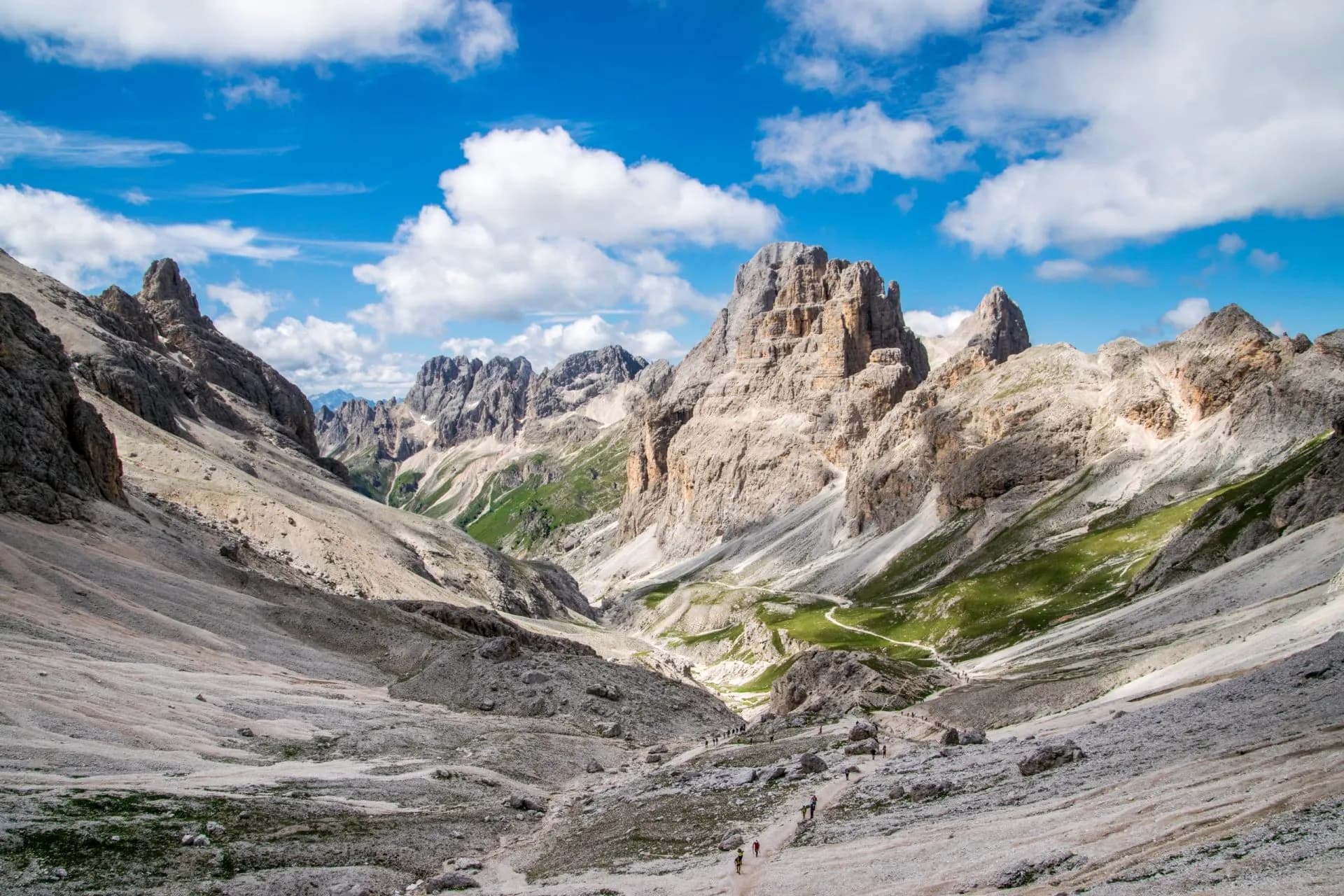 Hikers on rocky trail through steep limestone mountains under a blue sky with clouds in Rosengarten.