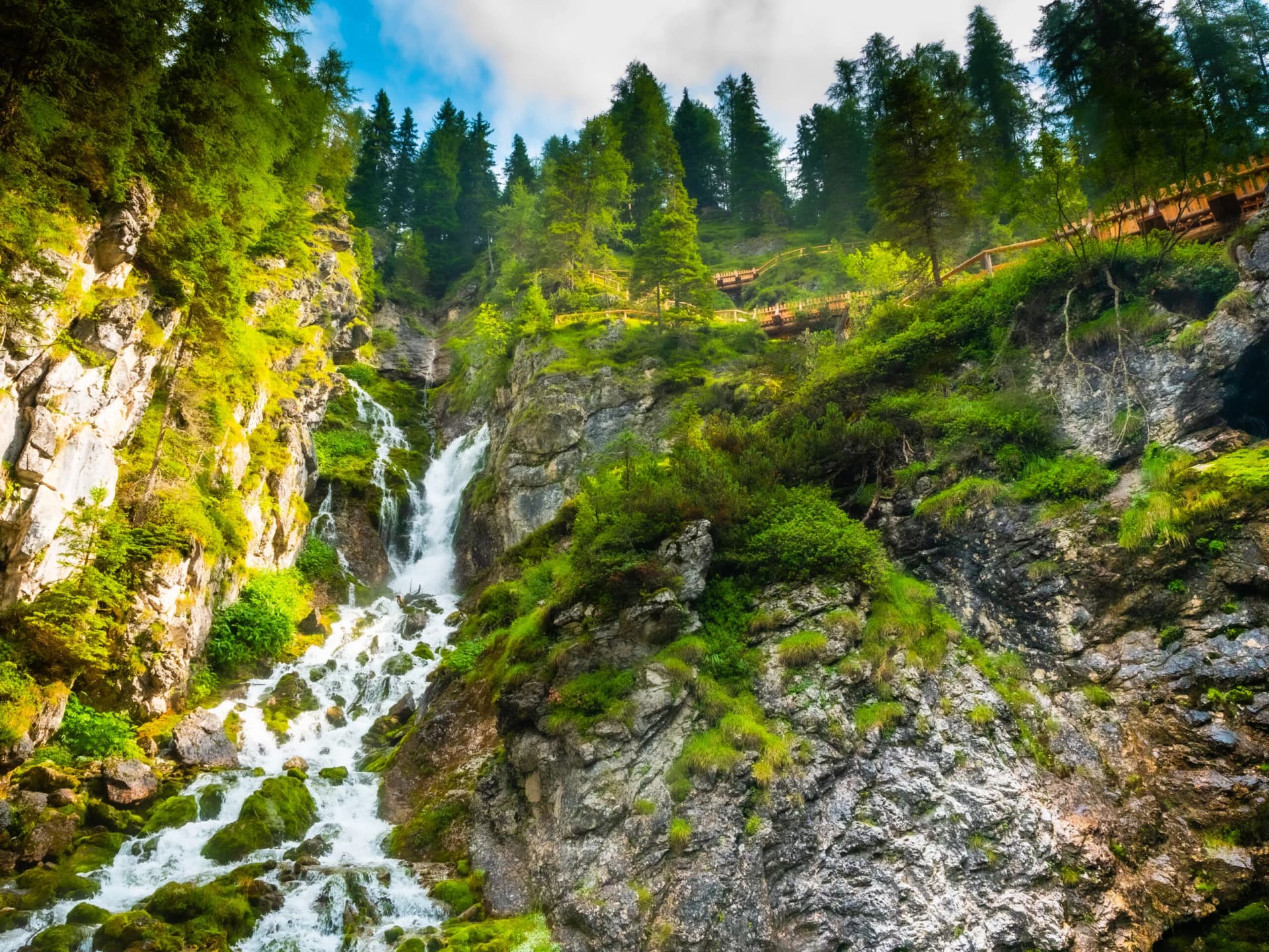 Vallesinella Waterfalls cascading down mossy rocks in the woods of Brenta Dolomites with wooden walkways.