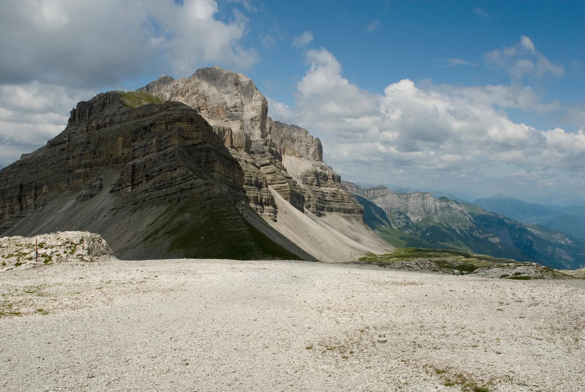 Rocky alpine plateau overlooking layered mountain slopes under a partly cloudy blue sky.