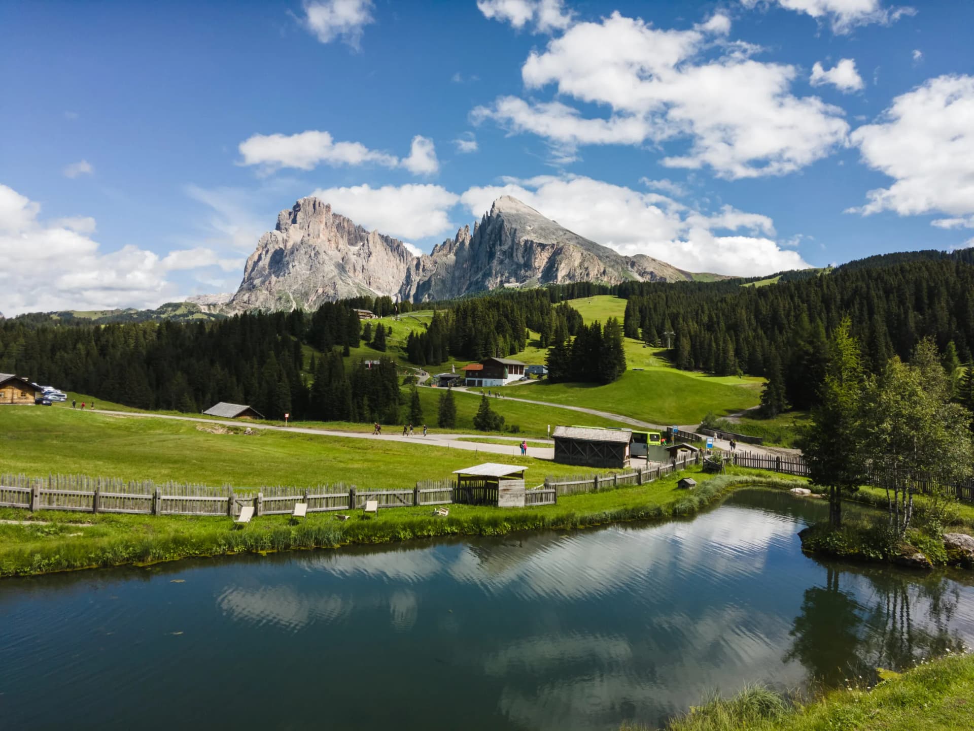 Pond reflecting clouds near grassy meadows, with Langkofel mountains in the background.