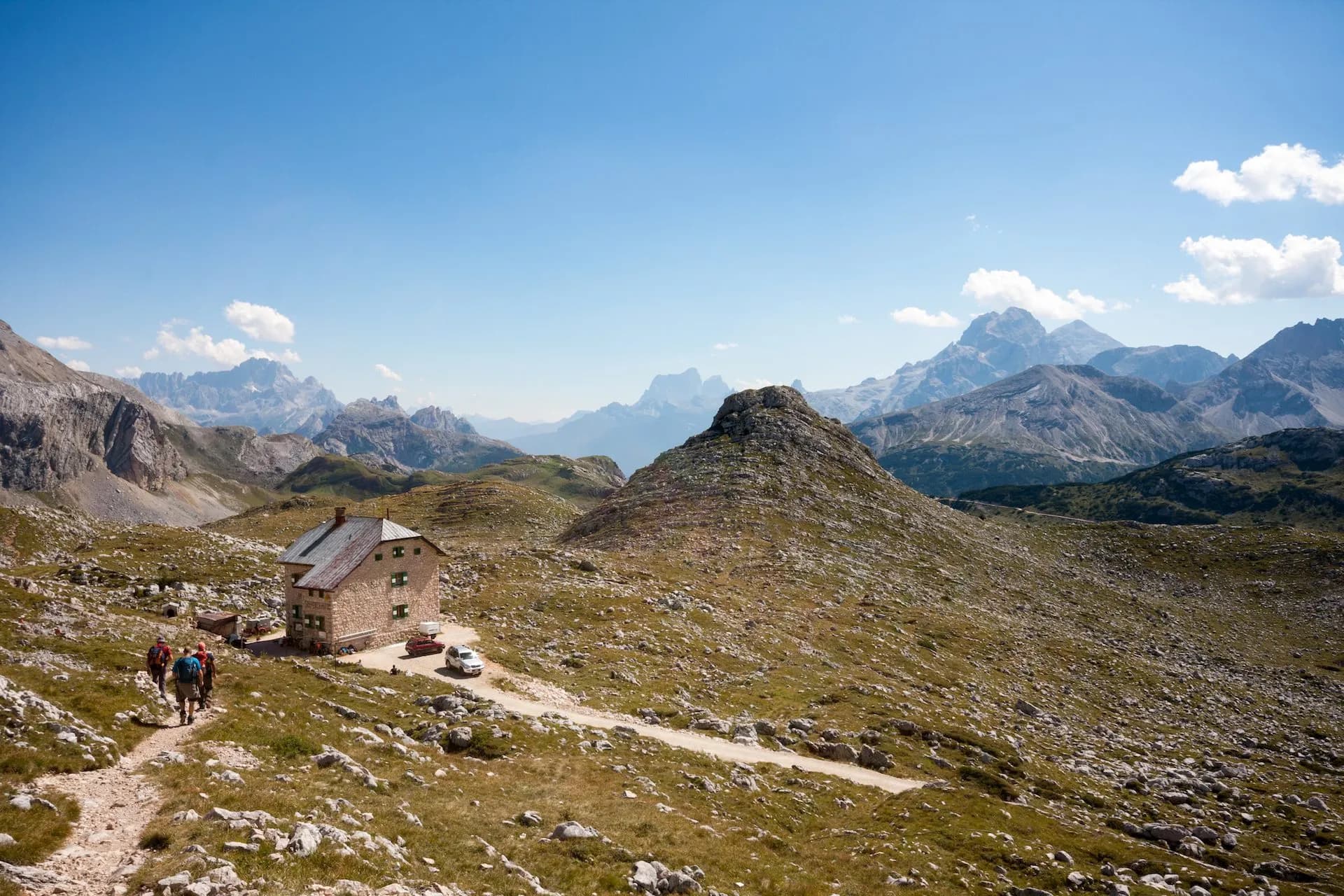 Hikers approach Rifugio Biella stone mountain hut surrounded by rocky alpine terrain under a blue sky.