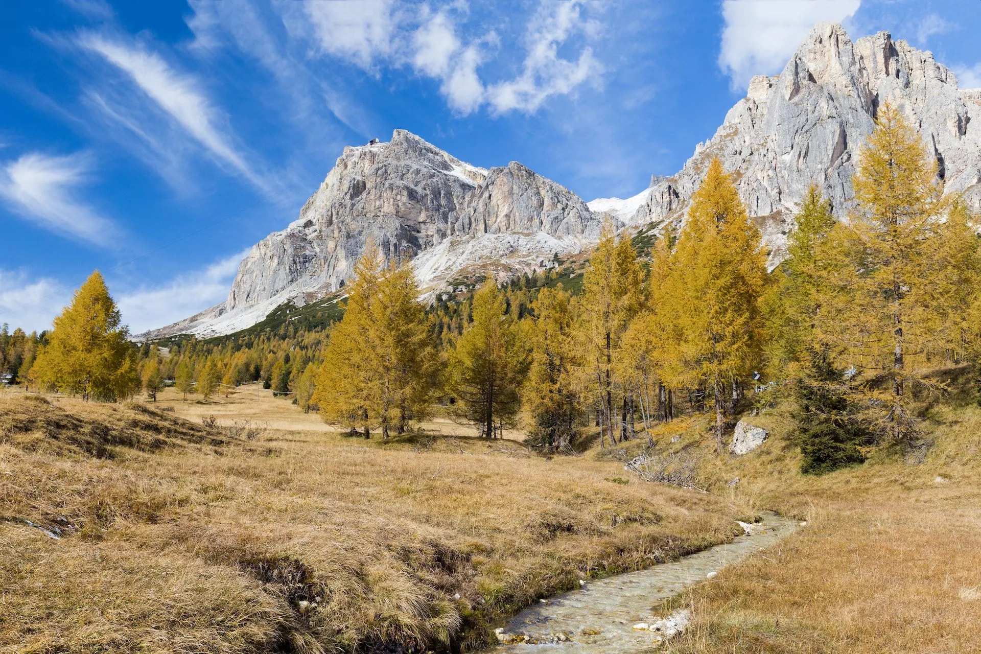 Stream flows through dry autumn meadow below rocky Falzarego mountains under blue sky.