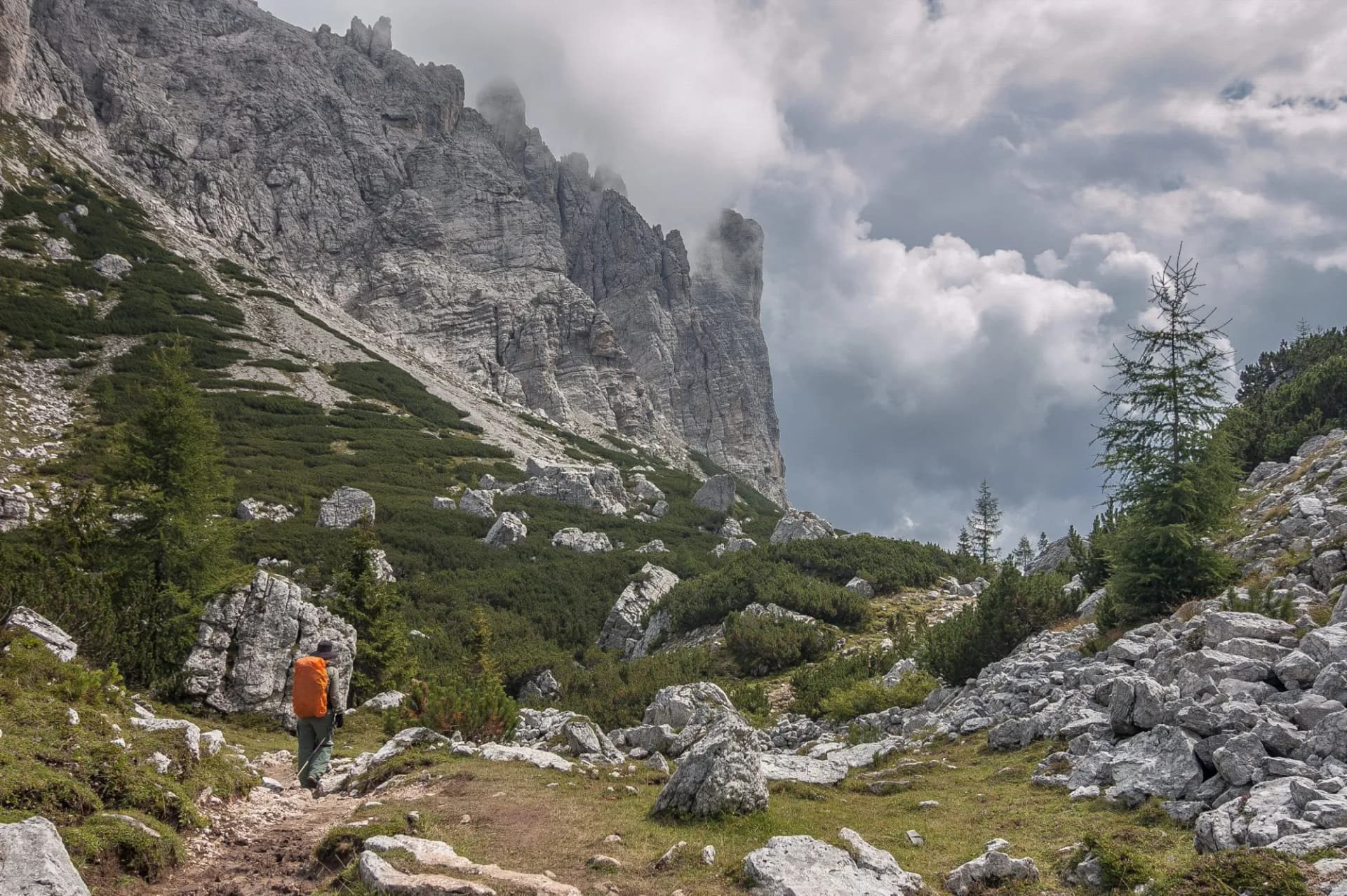 Hiker with orange backpack on rocky trail below sheer mountain cliffs under cloudy sky, Coldai to Vazzoler.