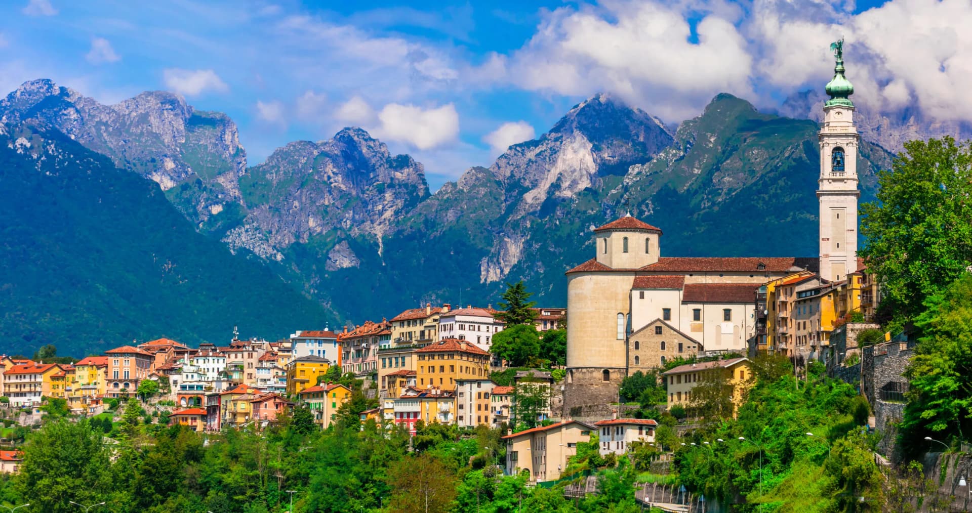 Belluno town with colorful buildings, church tower, and lush green slopes backed by Dolomite mountains.