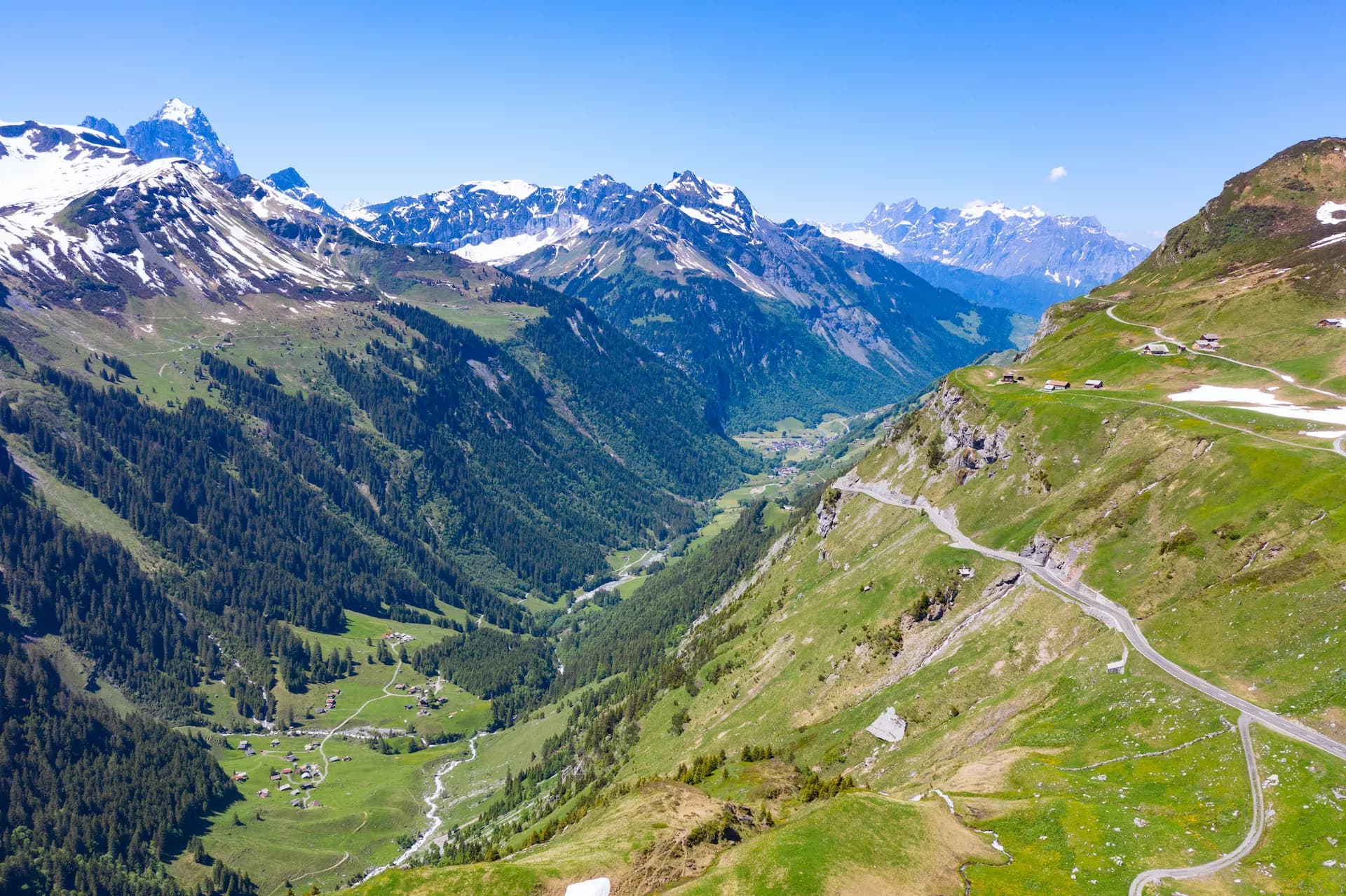 Winding mountain road connecting cantons Uri and Glarus over green slopes with snow-capped peaks.