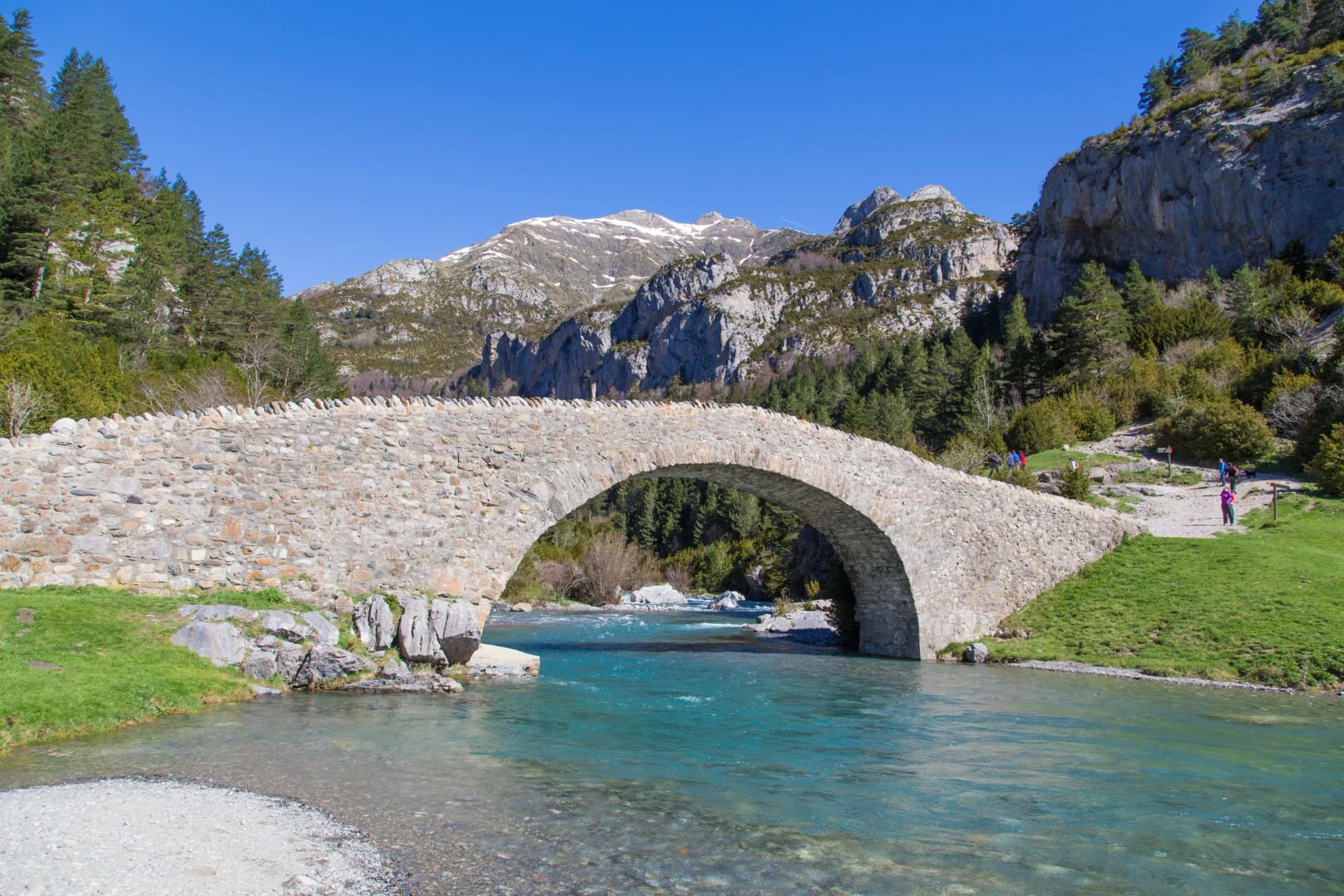 Stone arch bridge over turquoise river with hikers near snow-capped mountains.