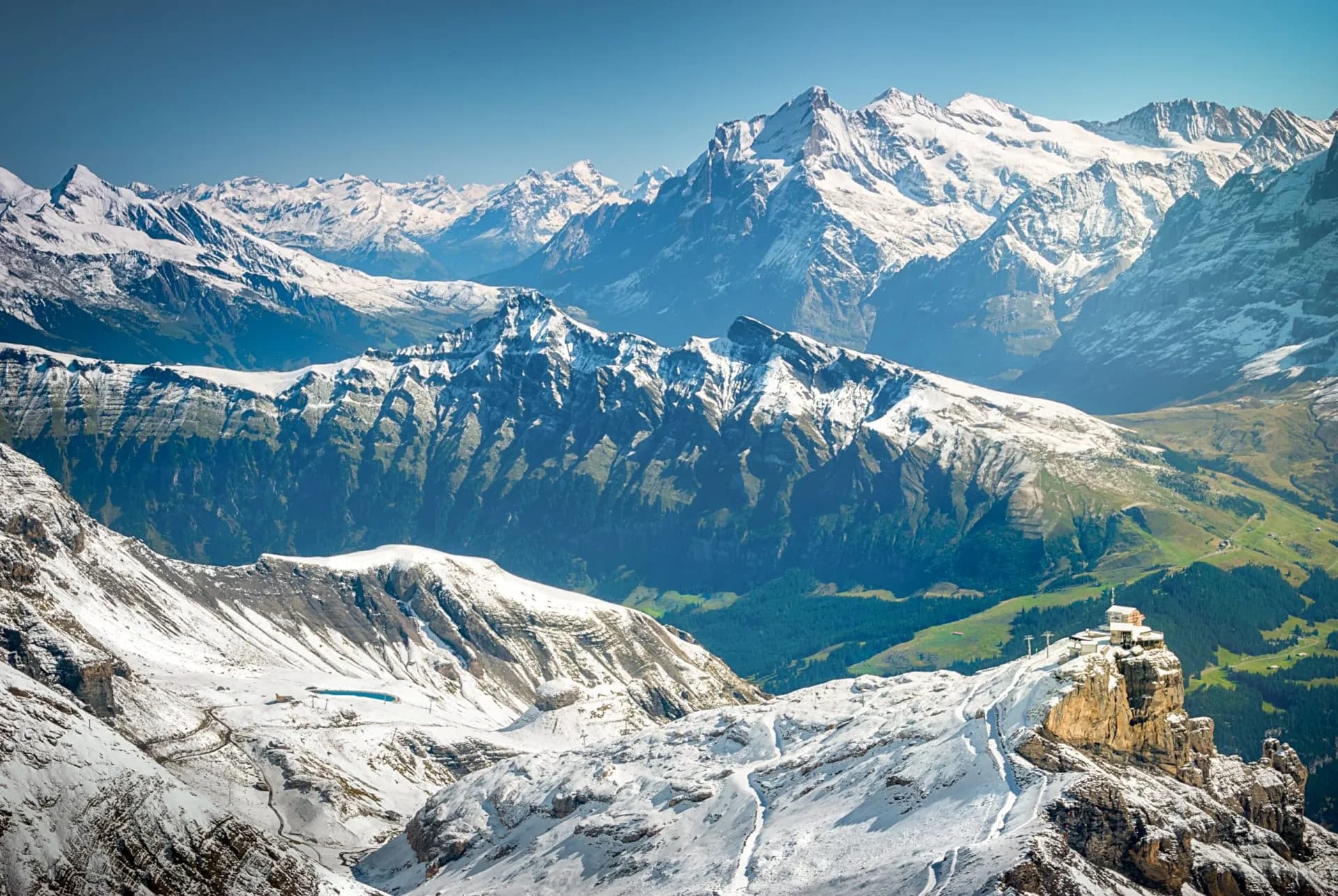 View of Jungfrau region mountains from Schilthorn with snow, green valleys, and cliffside building.